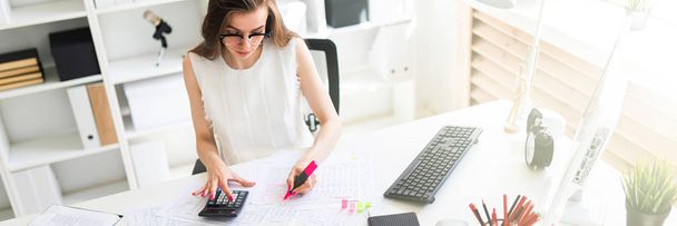 Una joven en la oficina lleva un marcador rosa en la mano y cuenta con una calculadora
. - Foto, imagen