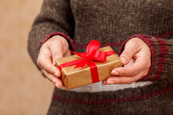 Woman in a sweater holding a gift box tied with a red ribbon in her hands. Shallow depth of field, Selective focus on the box. Concept of giving a gift on Valentine's Day or birthday. - Fotó, kép