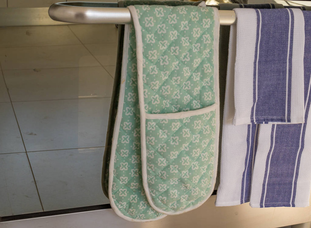 Μια πετσέτα πιάτο και ένα ζευγάρι γάντια φούρνου κρέμονται πάνω από τη ράγα της σόμπας σε μια εικόνα εγχώρια κουζίνα με χώρο αντίγραφο σε οριζόντια μορφή - Φωτογραφία, εικόνα