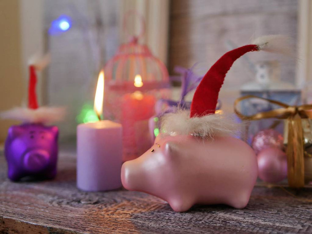 La composición decorativa estacional del juguete de Año Nuevo de cerdo en el sombrero rojo de Papá Noel, la iluminación de fiesta, las velas, las cajas con las bolitas, las linternas sobre la mesa de madera, el color rosado, el año del cerdo, la Navidad
 - Foto, imagen