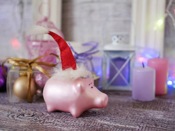Decorative seasonal composition of a New Year's pig toy in a red Santa hat, festive illumination, candles, boxes with balls, lanterns on a wooden table, pink color, the year of the pig, Christmas - Photo, Image