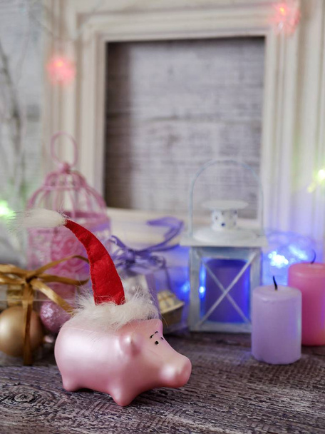  Декоративный сезонный состав игрушечных новогодних поросят в красных шляпах Санты, праздничное освещение, зажженные свечи, коробки с шариками, фонари на деревянном столе, розовый цвет, год свиньи, Рождество
 - Фото, изображение