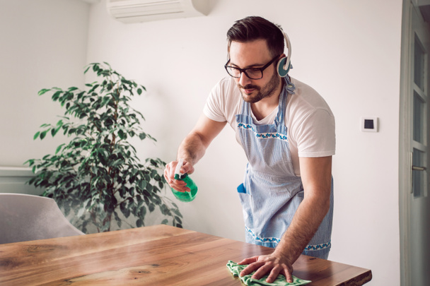 Young smiling man cleaning the table with a rag and spray bottle detergent - Foto, Imagen