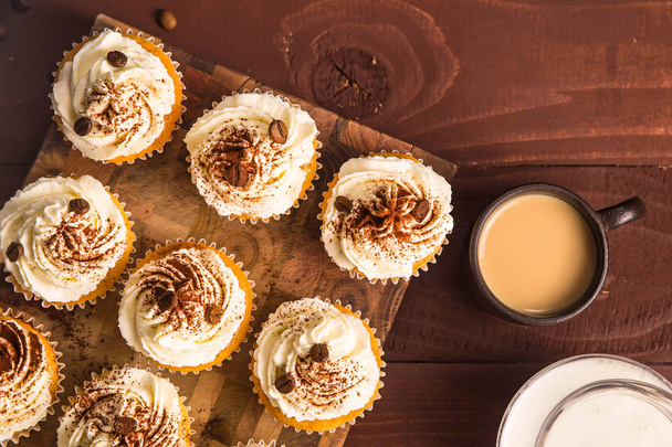 Tiramisu cupcakes decorated with cocoa powder and coffee grains on a wooden board next to jugs of milk, cream and a cup of coffee. Top view, flat lay - Photo, image