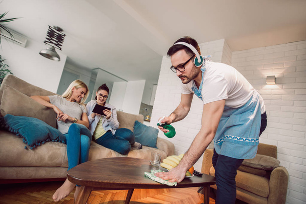 Ehemann Hauswirtschaft und Reinigungskonzept. ein Mann putzt das Haus, während Frauen auf dem Sofa tratschen - Foto, Bild