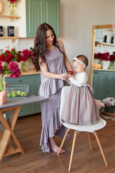 mamá y su hija en vestidos elegantes en la cocina, decorado con peonías
 - Foto, Imagen