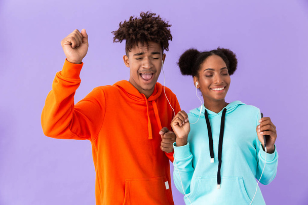Photo of african american couple in colorful clothes singing while listening to music together with earphones isolated over violet background - Photo, image