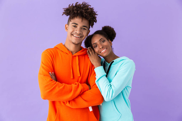 カラフルな服を着て立って一緒に紫色の背景に分離しながら笑みを浮かべて美しいアフリカ系アメリカ人カップルの写真 - 写真・画像