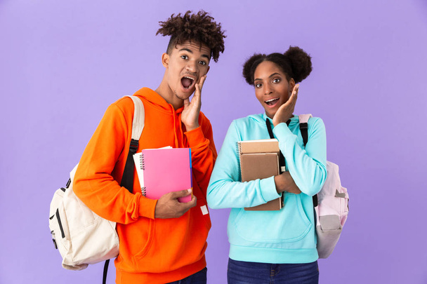 Photo of excited african american man and woman wearing backpacks holding exercise books isolated over violet background - Foto, Imagem