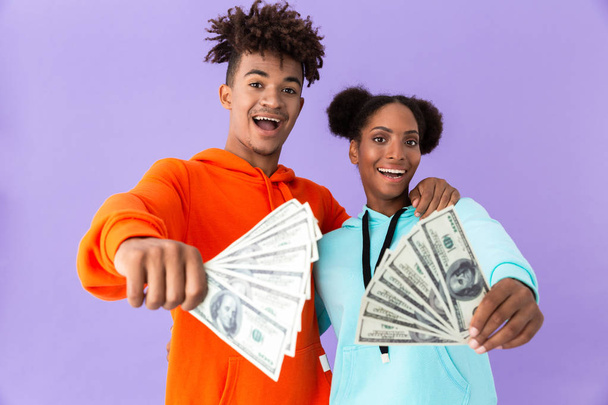 Photo of successful african american couple in colorful clothes holding fans of dollar money isolated over violet background - Valokuva, kuva