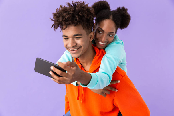 Photo of beautiful african american woman taking selfie while hugging brother from back isolated over violet background - 写真・画像