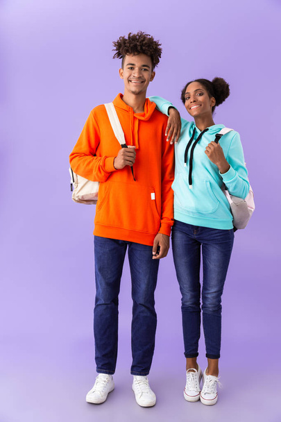 Full length photo of attractive african american brother and sister wearing backpacks hugging together isolated over violet background - Foto, Bild