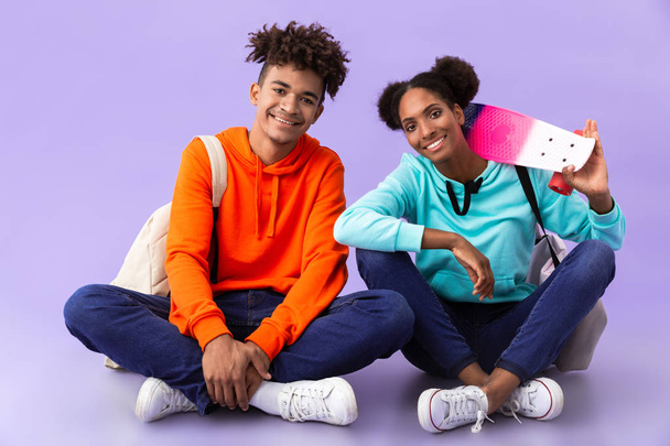 Image of teen african american people wearing backpacks sitting on floor with skateboard isolated over violet background - Foto, Imagem
