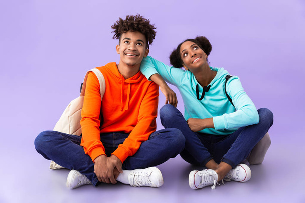Photo of teen african american man and woman wearing backpacks sitting on floor with legs crossed isolated over violet background - Foto, afbeelding
