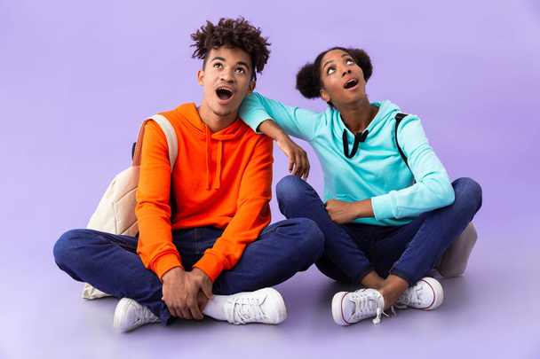Photo of cheerful african american man and woman wearing backpacks sitting on floor with legs crossed isolated over violet background - Φωτογραφία, εικόνα