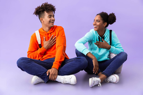 Photo of joyful african american man and woman wearing backpacks sitting on floor with legs crossed isolated over violet background - Foto, Imagen