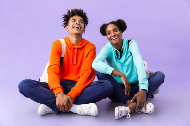 Photo of beautiful african american man and woman wearing backpacks sitting on floor with legs crossed isolated over violet background - Foto, imagen