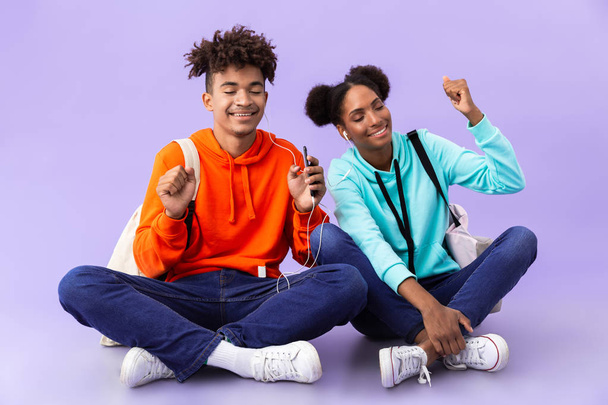 African american couple wearing backpacks using mobile phone and earphones while sitting on floor with legs crossed isolated over violet background - Foto, Bild