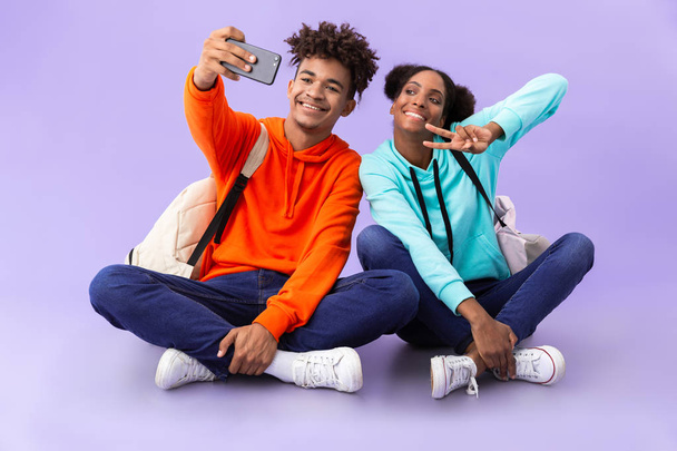 African american man and woman wearing backpacks taking selfie photo while sitting on floor with legs crossed isolated over violet background - Valokuva, kuva
