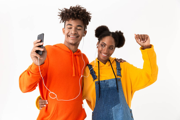 Foto van Afrikaanse Amerikaanse echtpaar in kleurrijke kleding dansen terwijl u luistert naar muziek samen met koptelefoon geïsoleerd op witte achtergrond - Foto, afbeelding
