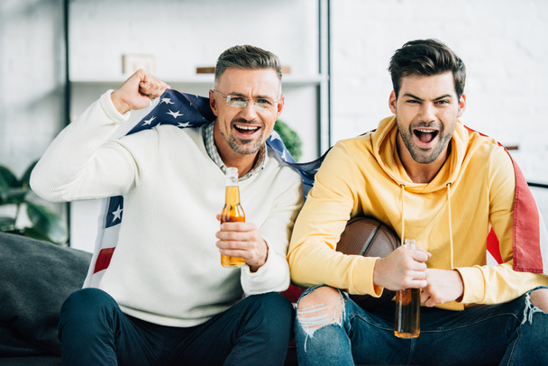 excited son and mature father wrapped in united states flag watching basketball game on weekend at home - Fotografie, Obrázek