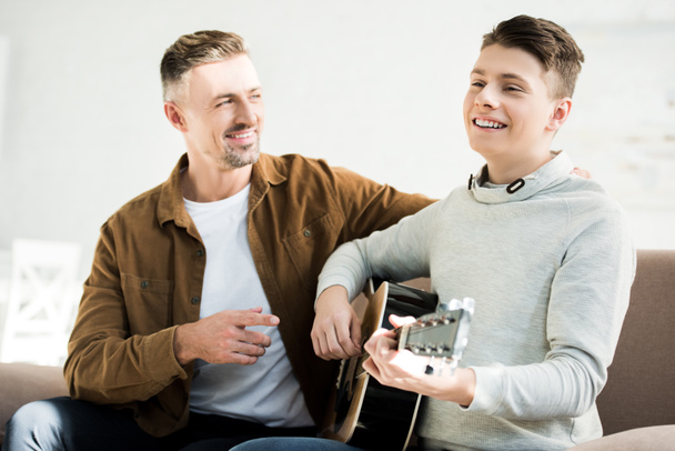 sonriente hijo adolescente tocando la guitarra acústica para el padre en casa - Foto, imagen