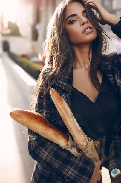 Vintage picture of a brunette woman in stylish black shirt and checkered grey jacket. Sexy fashion girl with long dark hair holding tasty french baguette in the hands. Warm autumn sunset colors. - Photo, Image