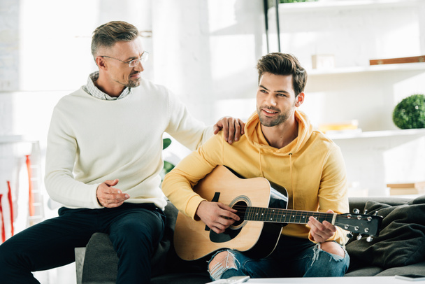 lachende zoon gitaarspelen voor volwassen vader op weekend thuis - Foto, afbeelding