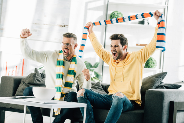 excited son and mature father with scarfs watching sport and screaming at home - Photo, Image