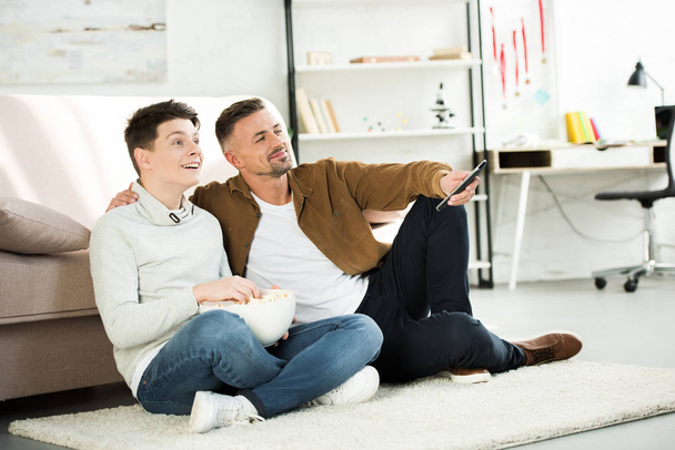 happy father and teen son watching tv and holding bowl of popcorn on floor at home - Foto, imagen