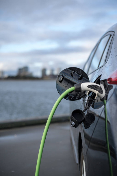 Φορτίζει το ηλεκτρικό αυτοκίνητο, με την πόλη και το νερό στο παρασκήνιο, πράσινο καλώδιο φόρτισης μαύρο αυτοκίνητο. Κοπεγχάγη - Φωτογραφία, εικόνα