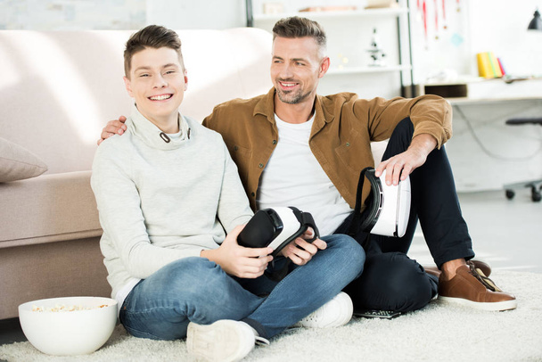 happy father and teen son holding virtual reality headsets at home - Foto, afbeelding