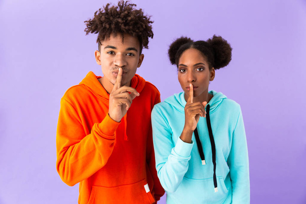 Photo of young african american guy and girl holding index fingers on lips meaning shh isolated over violet background - Foto, imagen