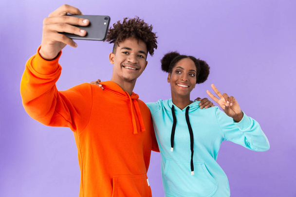 Foto van de gelukkige Afro-Amerikaanse paar dragen kleurrijke sweatshirts selfie nemen op smartphone geïsoleerd over violette achtergrond - Foto, afbeelding