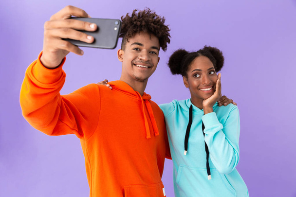 Photo of teenage african american couple wearing colorful sweatshirts taking selfie on smartphone isolated over violet background - Foto, imagen