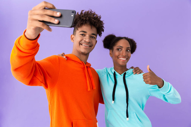 Photo of joyous african american couple wearing colorful sweatshirts taking selfie on smartphone isolated over violet background - 写真・画像