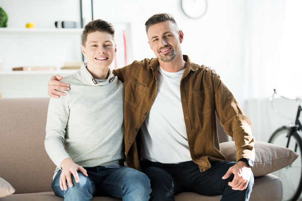 happy father and teen son sitting on sofa and looking at camera at home - Foto, Imagen