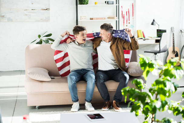 feliz padre e hijo adolescente envuelto en la bandera de los estados unidos sentado en el sofá y gritando durante el partido deportivo
 - Foto, imagen