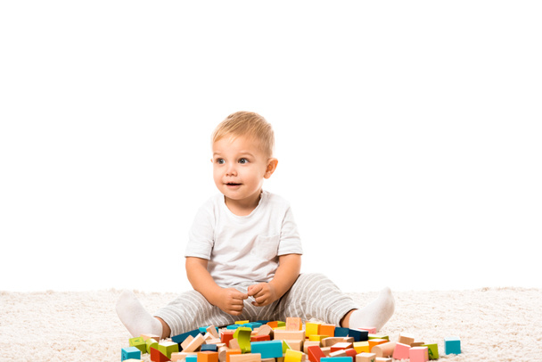 smiling toddler boy sitting on carpet and playing with multicolored wooden building blocks isolated on white - Фото, зображення
