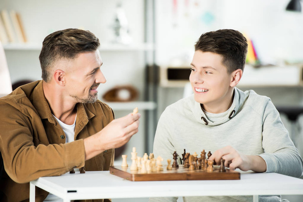 smiling father and teen son playing chess at home and looking at each other - Фото, зображення