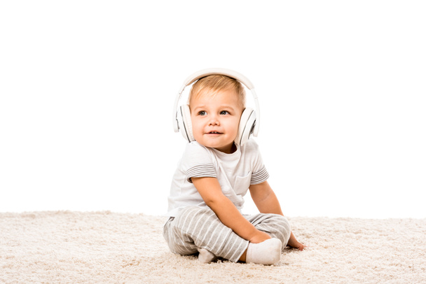 toddler boy sitting on carpet with headphones isolated on white - Photo, Image