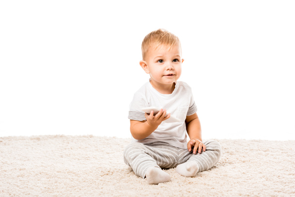 adorabile bambino ragazzo con smartphone seduto su tappeto isolato su bianco
 - Foto, immagini