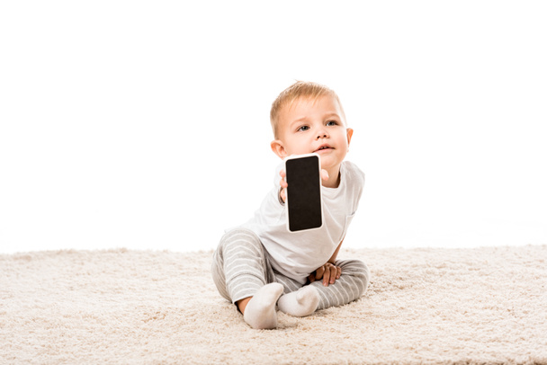 nice toddler boy sitting on carpet and showing smartphone screen isolated on white - Photo, Image