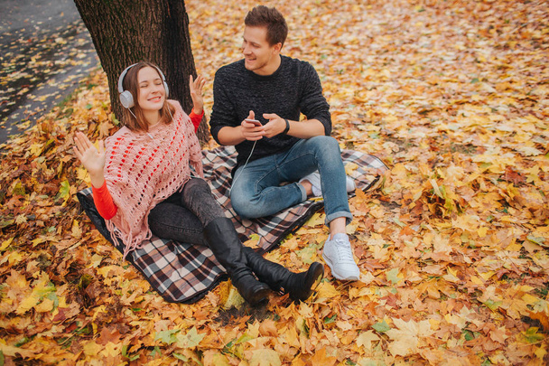 Young and positive young woman enjoys listening to music through headphones. She sits on blanket. Guy is besides her. He holds phone and look at woman. - Fotografie, Obrázek