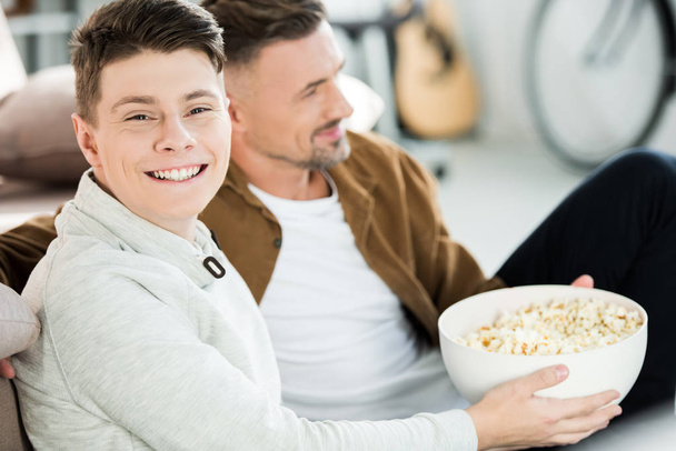 portrait de père et heureux fils adolescent regarder la télévision et tenant bol de maïs soufflé à la maison
 - Photo, image