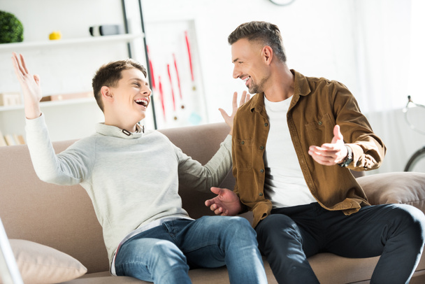 happy father and teen son sitting on sofa, gesturing and looking at each other - Photo, Image