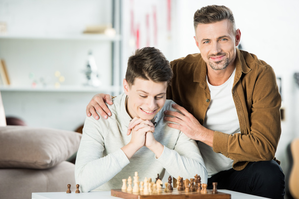 cheerful father hugging teen son while playing chess at home - Foto, afbeelding