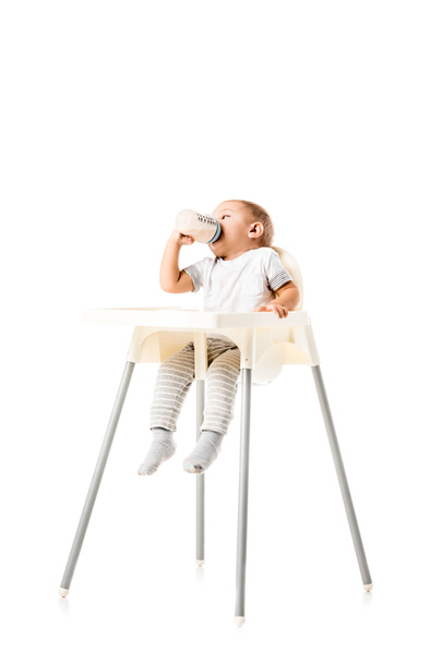 cute toddler boy drinking from baby bottle and sitting in highchair isolated on white - Photo, Image