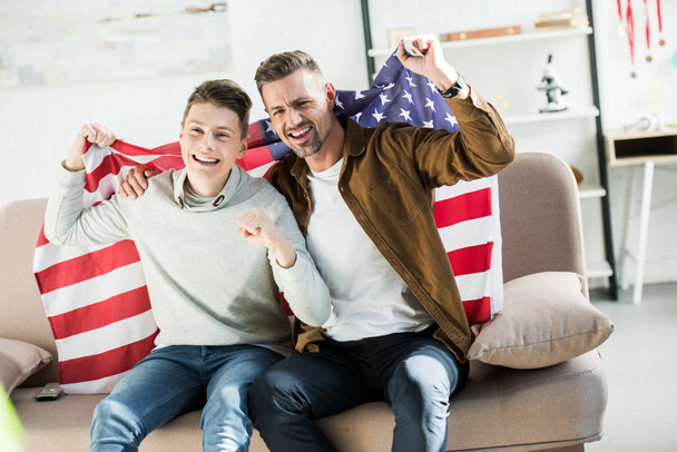 happy father and teen son wrapped in united states flag sitting on sofa and watching sport match - Photo, Image