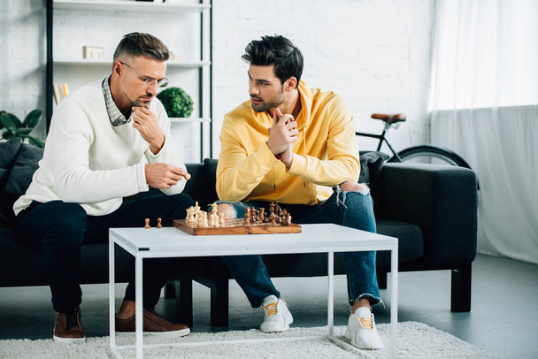 pensive son and mature father playing chess together on weekend at home - Фото, зображення